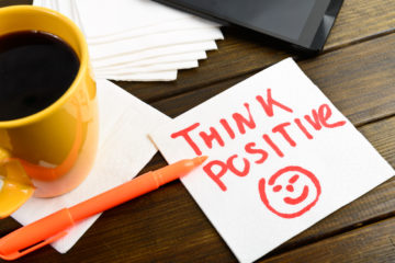 Think positive with a smiley face, writen on white napkin in orange pen, next to a cup of coffee pen on a wooden table