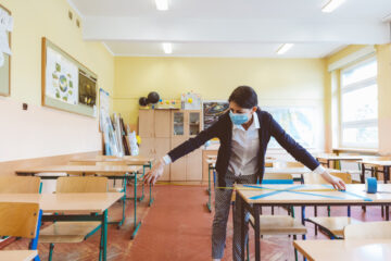 A teacher wearing a facemask measuring the distance between desks in a classroom.