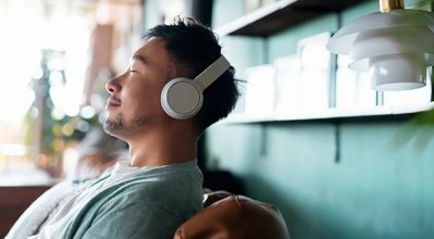 (alt="Young man with eyes closed, enjoying music over headphones while relaxing on the sofa at home”)